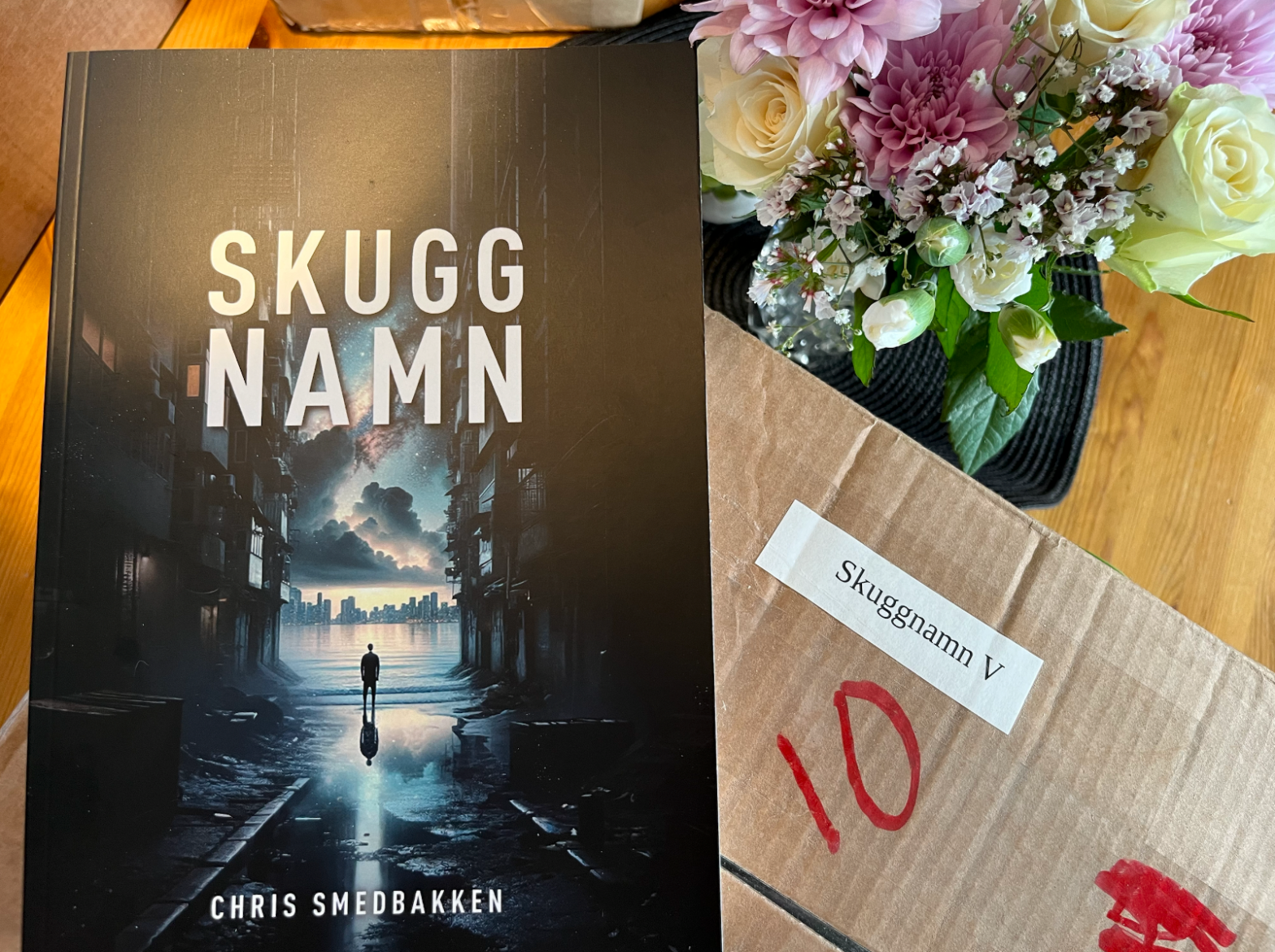 Boken Skuggnamn invid en blombukett och en kartong med samma namn på. Omslaget föreställer silhuetten av en person som står i slutet av en vattenfylld gränd och blickar ut mot en stad på andra sidan av en bukt.